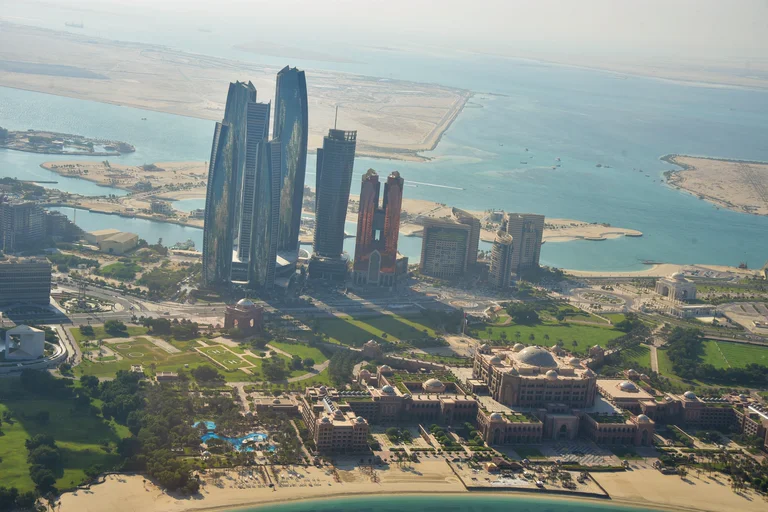 Curved glass towers rising behind Emirates Palace and its beach, with sandy islands in the turquoise sea beyond