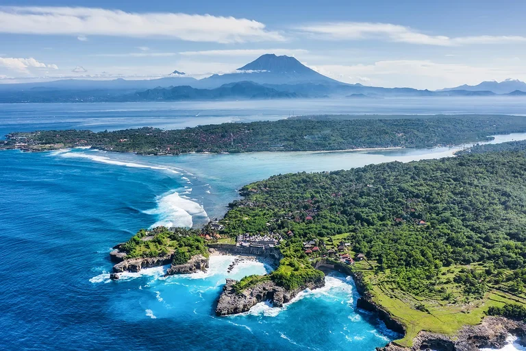 Green island headland and turquoise shallows off Bali, with a tall volcano on the horizon beyond the sea