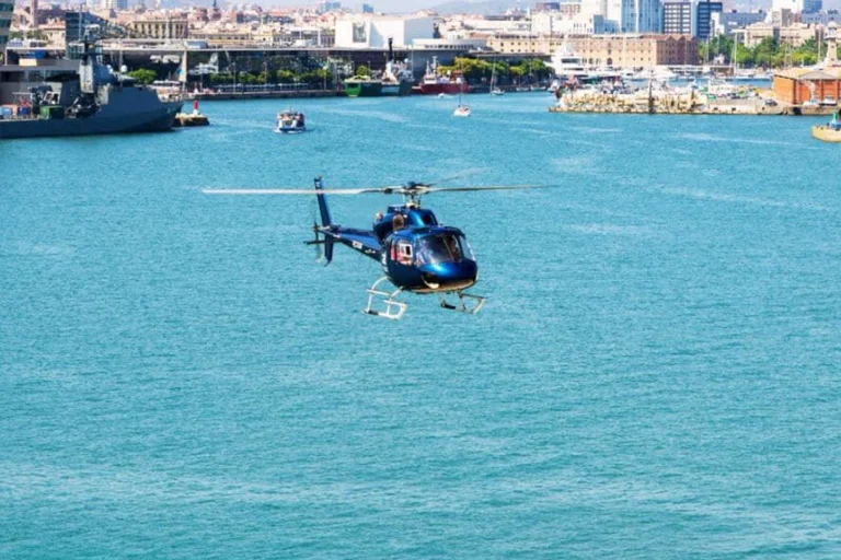 Blue helicopter flying low over turquoise water in the Port of Barcelona, with ships and the city waterfront behind.