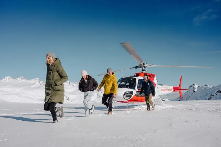 Guests walking on snow away from a red-and-white helicopter under blue sky at Fox Glacier.