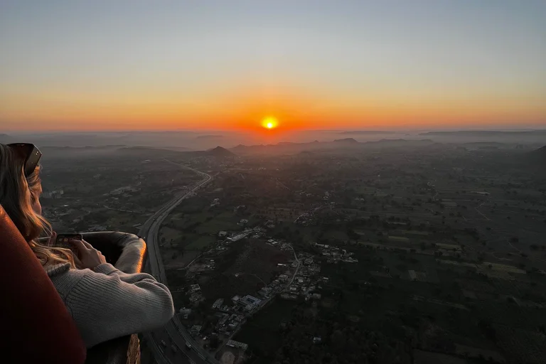 Passenger at a hot air balloon basket rim watching the sun rise over flat countryside, low hills and a curving highway