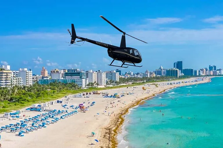 Black helicopter flying over South Beach, above rows of blue umbrellas, turquoise water and a line of hotels and towers