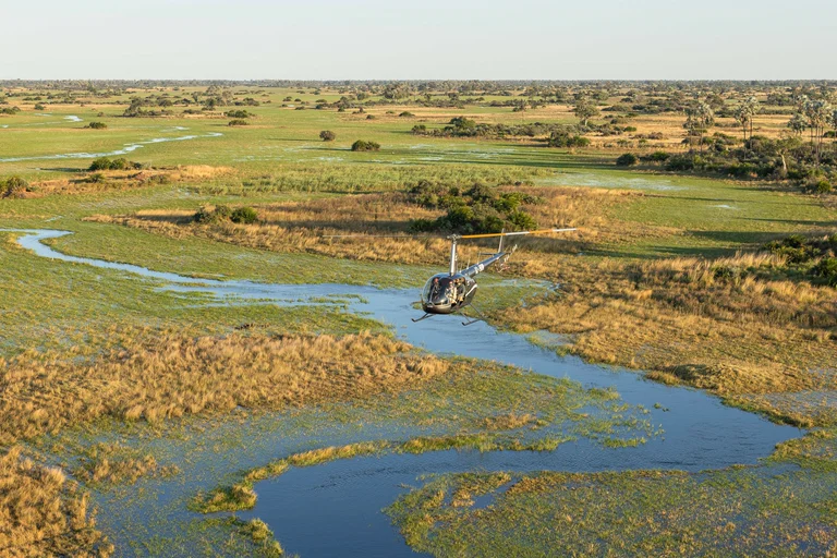Helicopter with passengers flying low over a winding blue channel through green floodplain