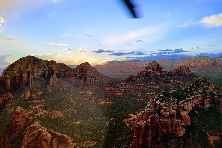 View from a helicopter over Sedona's red-rock buttes and green valleys at golden hour; a blurred rotor blade at the top, blue sky with scattered clouds.